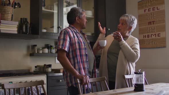Happy senior mixed race couple having coffee talking in kitchen alt