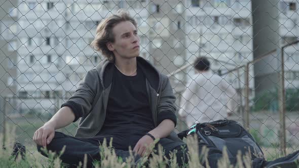 Upset Caucasian Teenager Sitting on Grass at Mesh Fence and Thinking. Sad Young Man Looking Back at alt