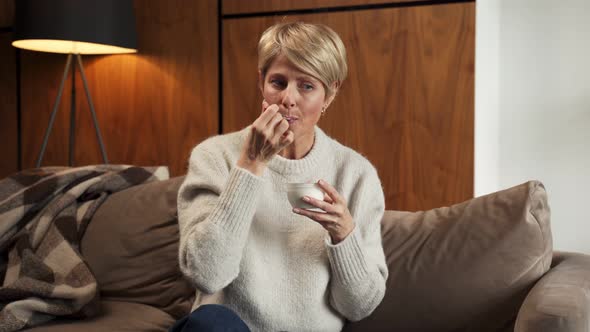 Portrait of a Middleaged Woman Who Eats Yogurt Sitting on the Couch at Home alt