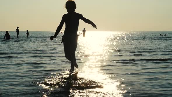 Child Runs In The Sea Water In Sun Flare, Kid Silhouette Running In The Sea. Sun Flare