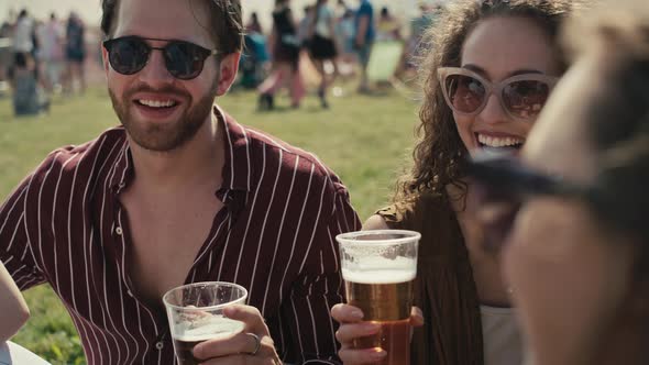 Group of friends sitting on grass together at music festival and drinking beer while chatting. Shot alt