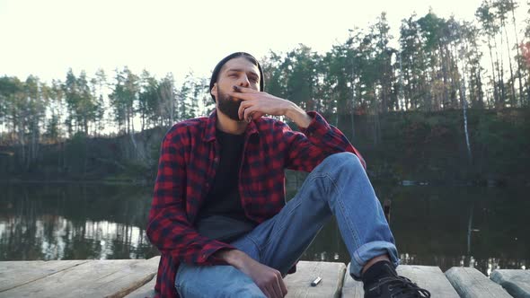 Bearded Yung Man Sit Near Amazing Lake and Smoke Cigar alt