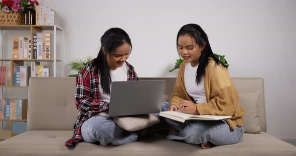 Twin girls reading a book and looking laptop while sitting on couch alt