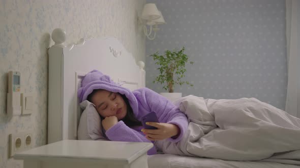 20s Asian Woman in Purple Pajamas Lying in Bed Using Mobile Phone