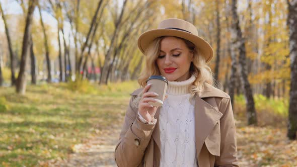 Young Woman in a Brown Coat and Hat Walks in the Autumn Park and Drinks Coffee alt