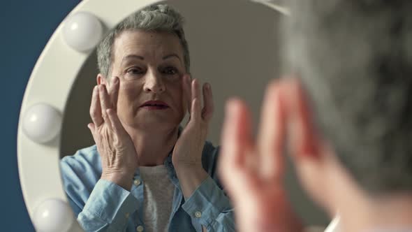 An Elderly Graying Woman Carefully Examines Her Reflection in the Mirror Lightly Touching the alt