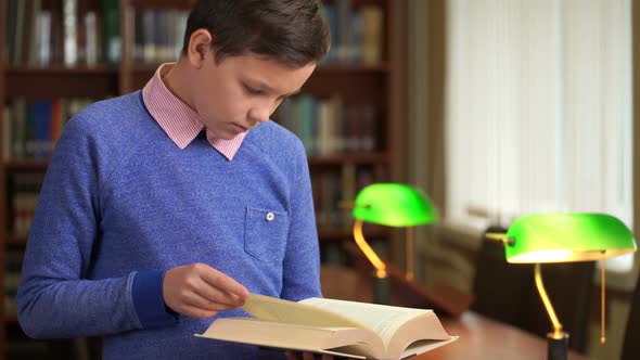 Portrait Shot of the Cute Schoolboy and Standing Near the Bookshelf in the Library and Reading alt