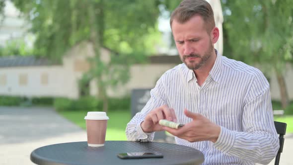 Middle Aged Man Feeling Sad While Counting Dollars in Outdoor Cafe alt