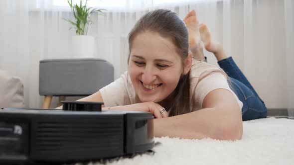 Portrait of Happy Smiling Woman Lying on Carpet and Looking on Working Robot Vacuum Cleaner alt