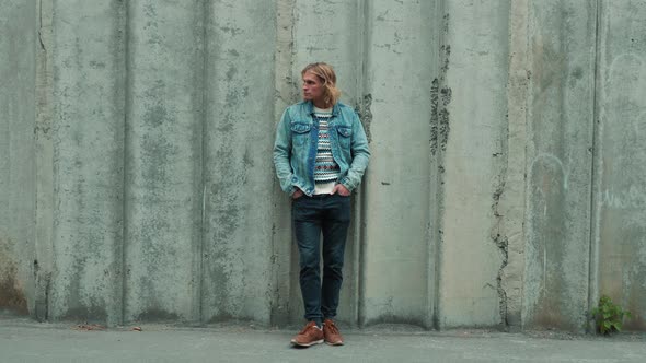 Fashionable young attractive man with blue eyes, long blond curly hair in blue denim jacket standing