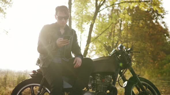 Young Attractive Man in Leather Jacket and Stylish Sunglasses is Standing By His Bike on the Country alt