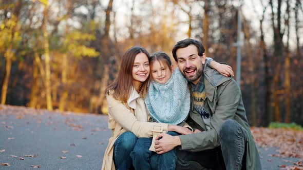 Young Smiling Mom Dad and Little Daughter are Hugging While Sitting Squatting in Autumn Park alt