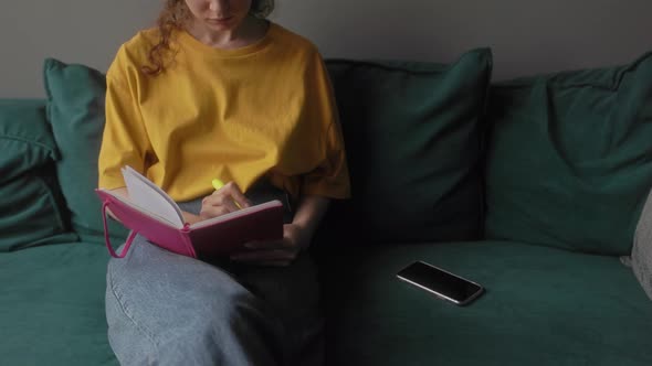 The Girl Makes Notes in a Notebook Sitting on a Bright Sofa