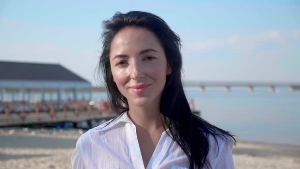 Portrait of Young Brunette Woman Looking at Camera Standing on the Beach with a Sea on Background alt