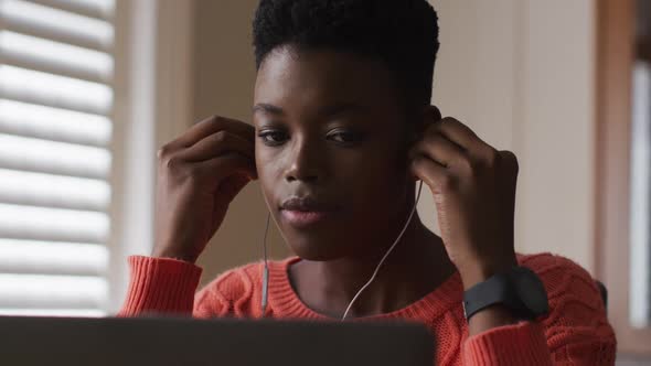 African american woman wearing earphones using laptop while working from home alt
