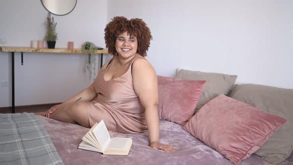 Beautiful Curvy Plus Size African Black Woman Afro Hair Lying on Bed in Silk Powder Pink Dress Cozy