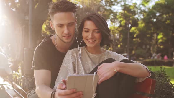 Young Beautiful Couple Smiling Speaking Looking at Tablet Sitting in City Park alt