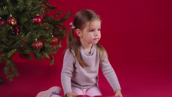 Little Cute Girl Unpacks Christmas Gift Sitting Near Xmas Tree, New Year's Eve