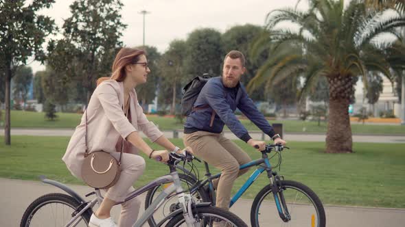 Cheerful Male and Female Bicyclists Are Chatting Happily in Walking in City Park alt