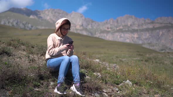 A Young Woman in a Sweatshirt Sits with a Phone in Her Hands Against a Background of Mountains. alt