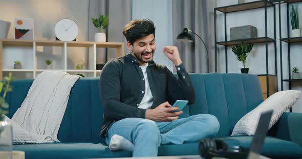 Guy Sitting on the Couch at Home and Getting Good news on Smartphone and Raising his Hands alt