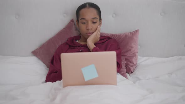 Portrait of Young Overworking African American Woman Lying in Bed Looking at Laptop Screen Closing alt