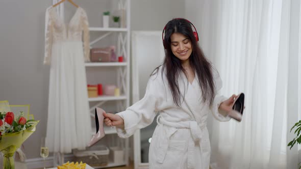 Joyful Happy Middle Eastern Woman in Headphones Dancing with Wedding Shoes in Living Room alt