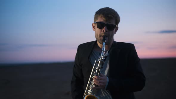 Bearded Man in Black Jacket and Glasses Play Saxophone Outdoors in Mountains on Plain at Dusk at alt