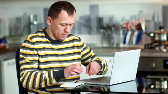 Man freelancer working on a laptop at home sitting at the table, background cuisine