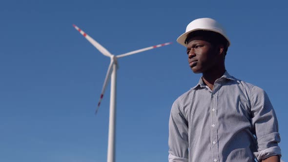 An AfricanAmerican Electrician in a Helmet Stands Against the Backdrop of a Windmill at an Air Power alt