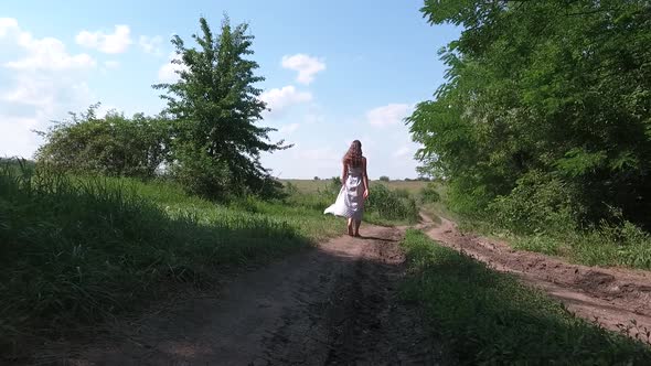 A Girl with Loose Hair in a Dress with Blue Polka Dots Walks Barefoot Into the Distance Along a Dirt