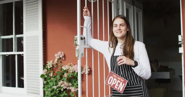 Smiling caucasian waitress standing in door, holding open sign, looking at camera alt