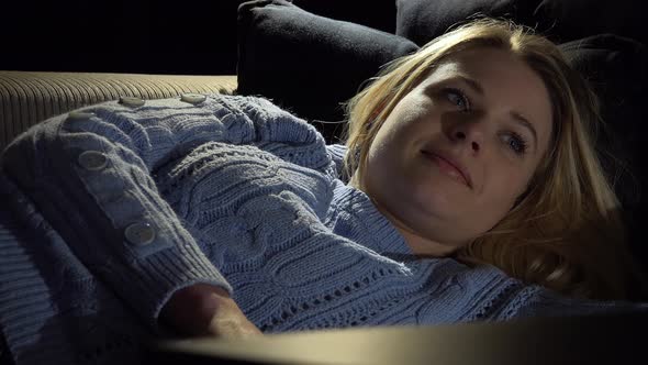 A Young Beautiful Woman Lies on a Couch in a Dark Room and Watches a TV - Closeup on the Woman alt