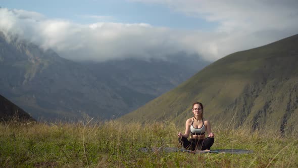 Young Woman in Tracksuit Practices Yoga Performing Lotus Position in the Mountains. alt