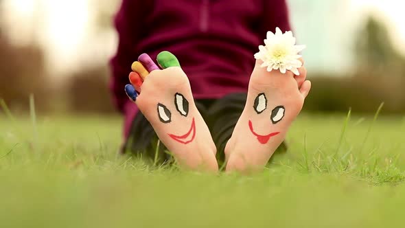 Little Child Girl Sits on Grass with Painted Feet with Funny Comic Faces alt