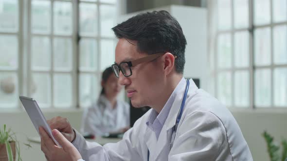 Asian Man Doctor Is Using Tablet While Work With Desktop Computer In Workplace. Medical Concept alt