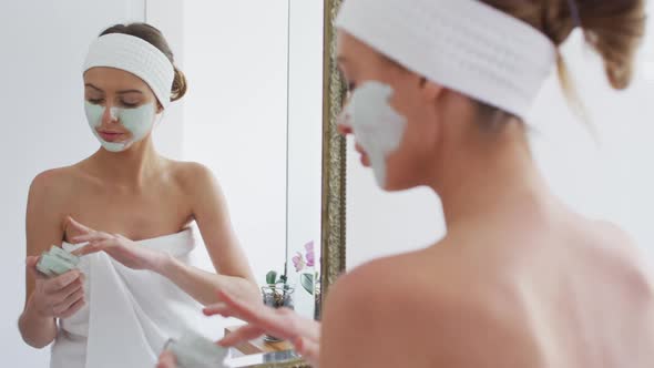 Woman applying face mask while looking in the mirror in bathroom alt