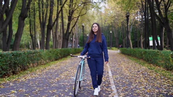Woman in Sport Costume with Bike Walking in Autumn Park. Happy Traveler Girl Biking in Park on Sunny