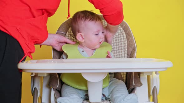 Mom puts on a bib toddler baby on a high chair for children, studio yellow background