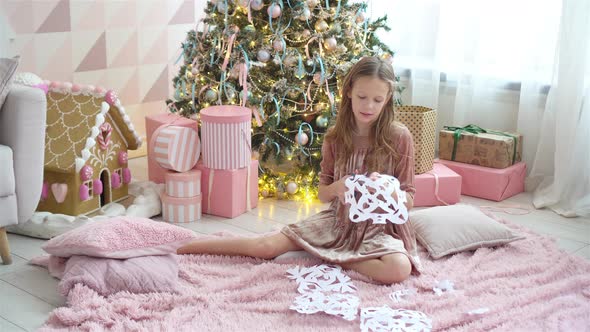 Adorable Little Girl Sitting Near the Tree and Making Paper Snowflakes alt