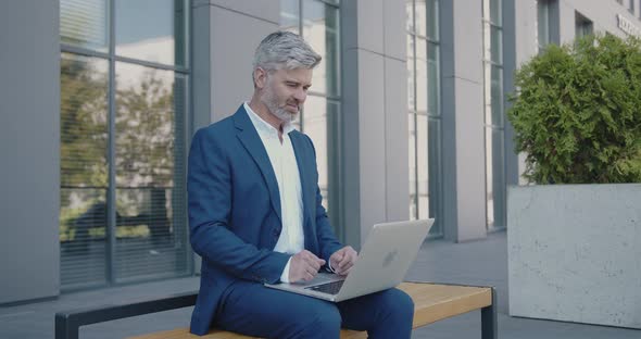 Mature Business Man Sitting on Bench and Working with Laptop in City Street alt