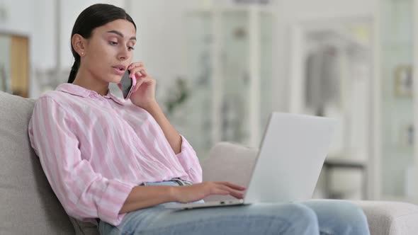 Serious Latin Woman with Laptop Talking on Smartphone at Home alt