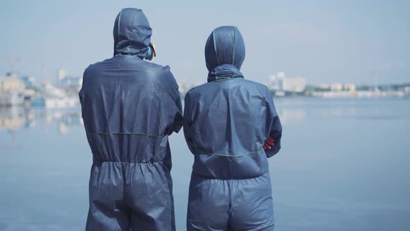 Back View of Two People in Biohazard Suits Standing at Riverbank and Looking at Blurred City on the alt