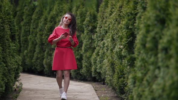 Stylish Woman in Red Dress is Walking in Green Alley in Park and Talking By Mobile Phone alt