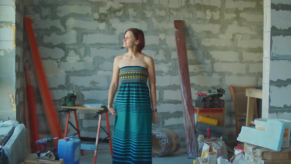 Contented Young Woman in Dress Stands in the Middle of a New Apartment Looking Forward alt