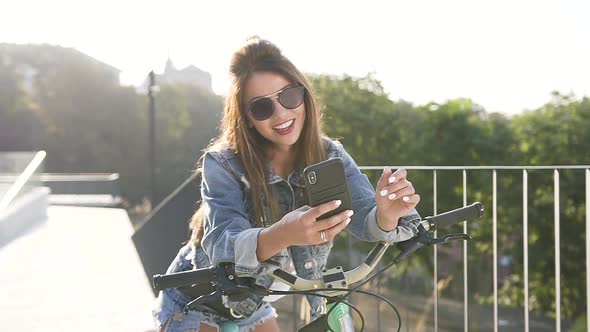 Young Woman in Sunglasses, Dressed in Jeans Clothing Walking with a Bicycle at the Park alt