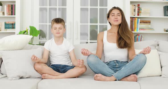 Goodlooking Positive Balanced Young Woman and Her 8Aged Son Which Meditating on Soft White Sofa in