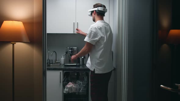 Young Man in Virtual Reality Goggles Unload Dishwashing Machine in Small Kitchen alt