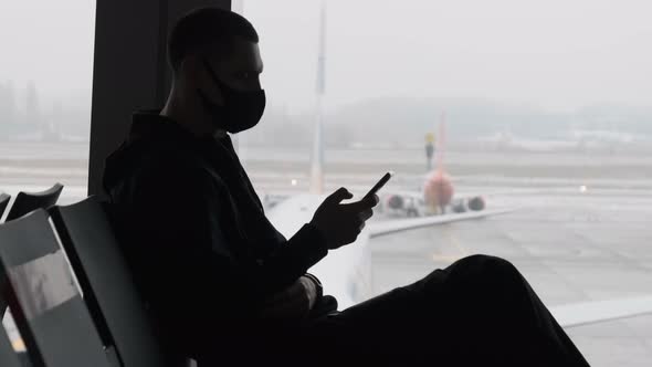 Masked Young Man in Airport Waiting Room Sits and Using a Smartphone By Window alt