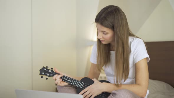 Young Woman Learning How To Play Ukulele on Laptop Computer Monitor, Watching Online Tutorial at